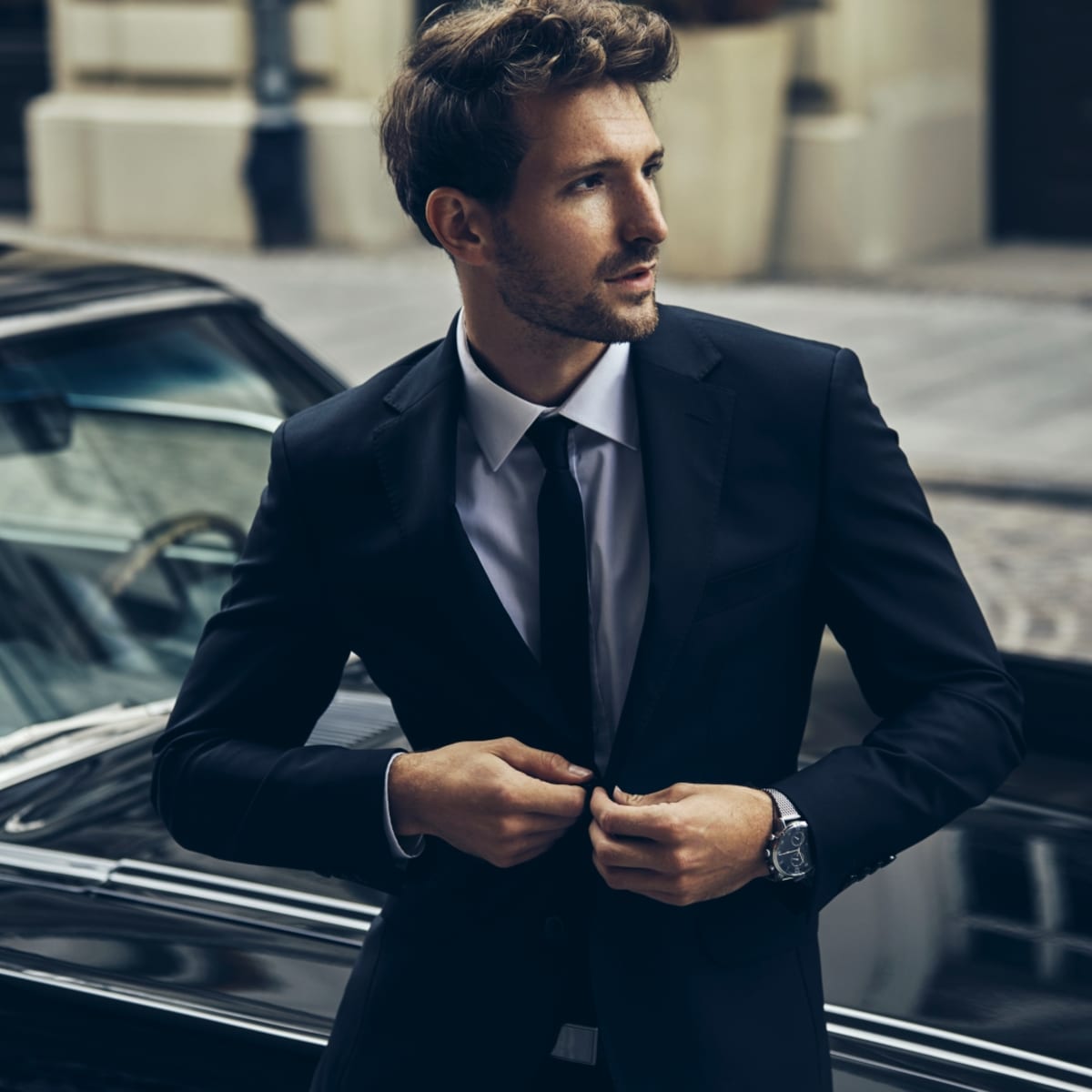 Wrist watch worn by a classique and young enterprenuer standing in front of a branded car who is putting a button on his coat in Laval.
