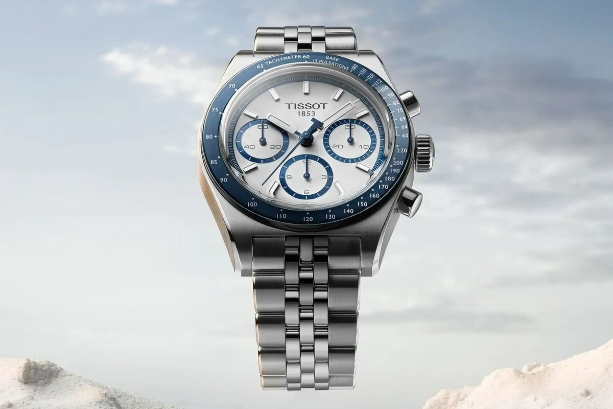 A Tissot 1853 chronograph watch with a silver stainless steel case and bracelet, featuring a white dial with blue subdials and a blue tachymeter bezel, set against a soft, cloudy sky background.