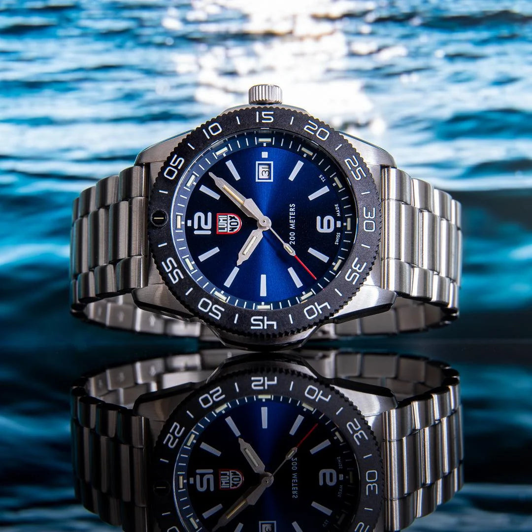 A stainless steel Luminox brand wristwatch with a blue dial and matching blue bezel, shown prominently over a reflective blue water surface. The watch has a silver metal bracelet and a black rotating bezel with white numerals. Shop sports watches at Bijouterie Caprices - Laval