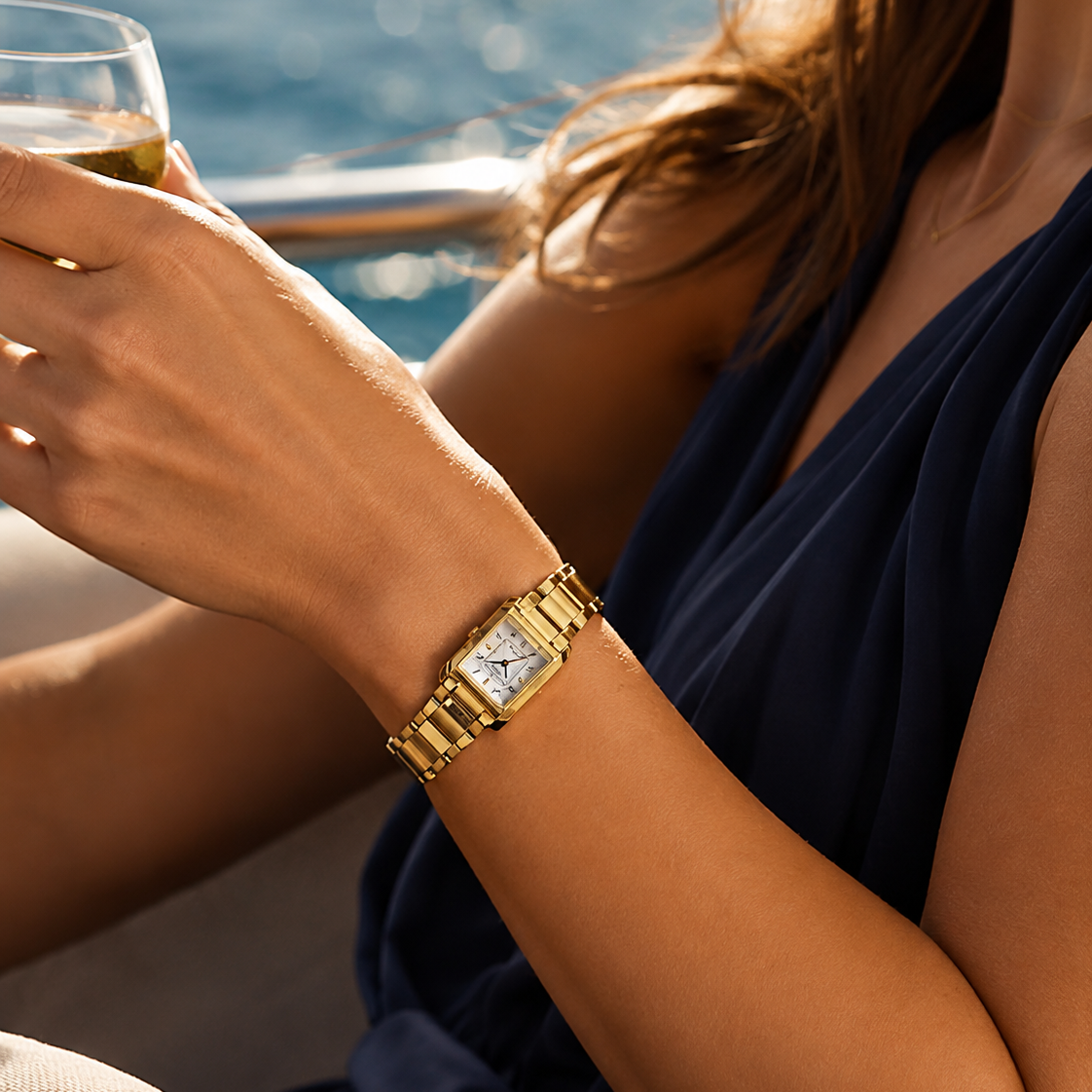 A woman in a navy blue dress wearing a gold-tone rectangular watch with a white dial, holding a glass of white wine by the water. Click to shop Womens luxury watches at Bijouterie Caprices - Laval